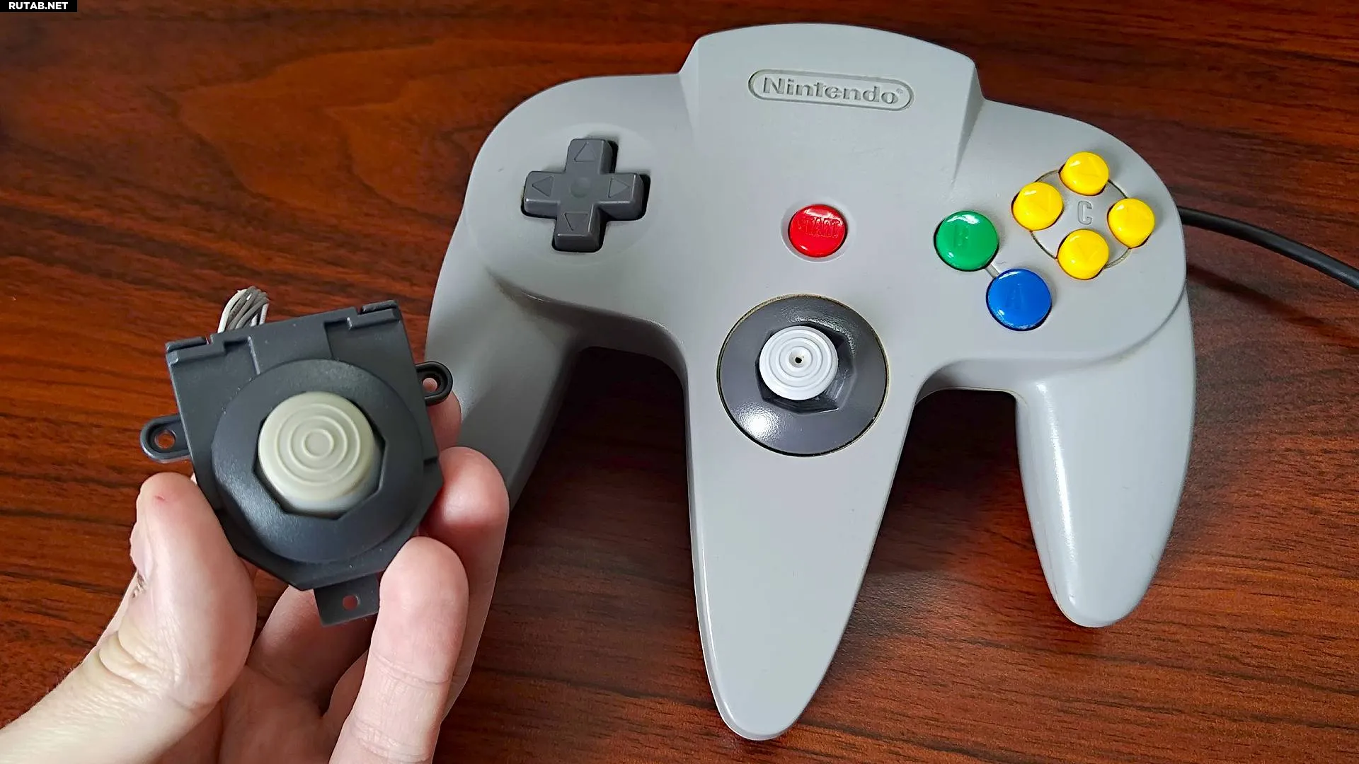 Рука держит стик Hyperkin на эффекте Холла рядом с серым контроллером Nintendo 64 на деревянном столе.